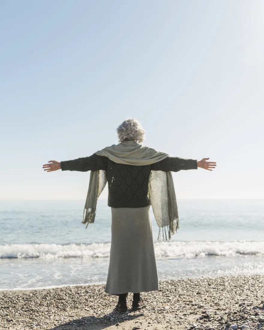 An older woman standing at the shoreline in the sun, arms open to the sea